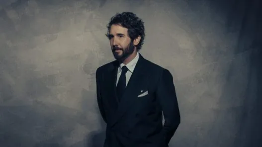 Man in a dark double-breasted suit and tie stands against a textured gray wall, looking to the left.