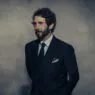 Man in a dark double-breasted suit and tie stands against a textured gray wall, looking to the left.