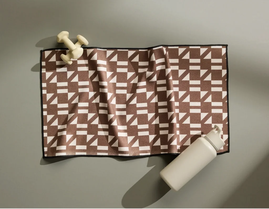 - Groovevolt Brown and white geometric patterned exercise mat on a gray floor, with a white water bottle nearby and light dumbbells at the edge.