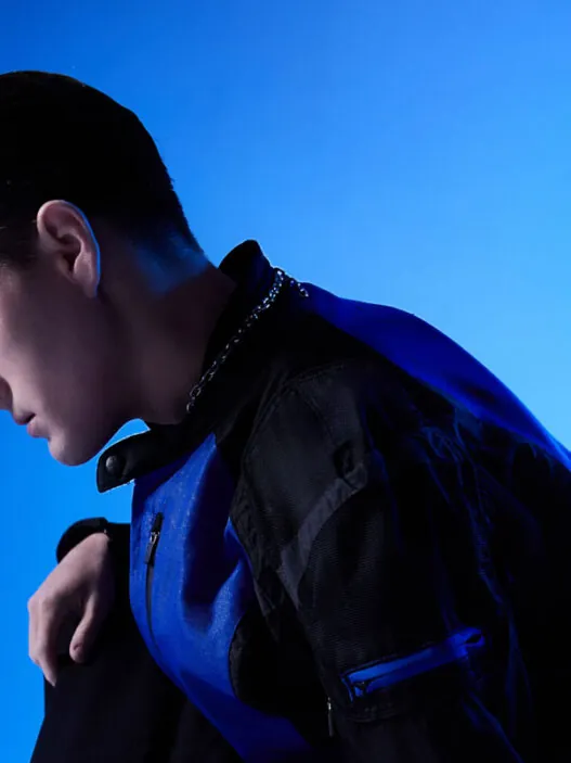 Person in a blue-and-black outfit kneels on a light floor, head bowed, left hand resting near the ground; dramatic blue gradient backdrop.