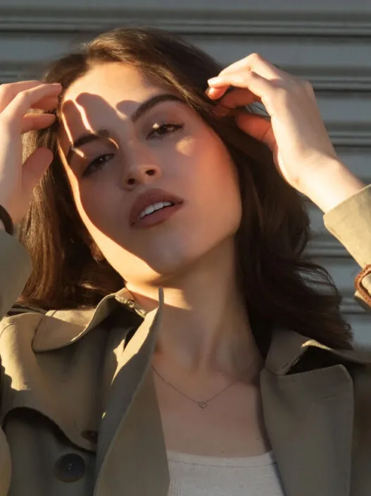 Young woman in a beige trench coat and white tank top, adjusting her hair in front of a corrugated metal backdrop.