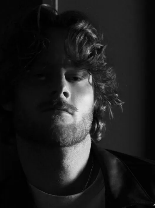 Black-and-white portrait of a man with wavy hair and a light beard, leaning against a wall in a leather jacket and casual shirt, looking slightly downward toward the camera.