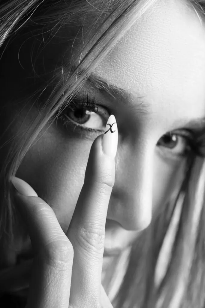 - Groovevolt Close-up of a woman's eye with a finger in the foreground, the fingernail showing a black 'x' mark near the eye.