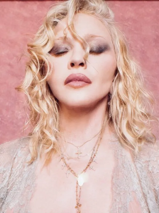 Close-up portrait of a blonde woman with wavy hair, eyes closed, wearing layered cross necklaces against a pink backdrop.