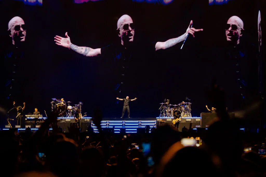 Lead singer with arms outstretched on a dark concert stage; band performs in the background and a large video screen shows his image.