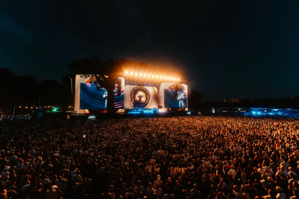 Massive outdoor concert crowd at night watching a brightly lit stage with circular LED visuals.