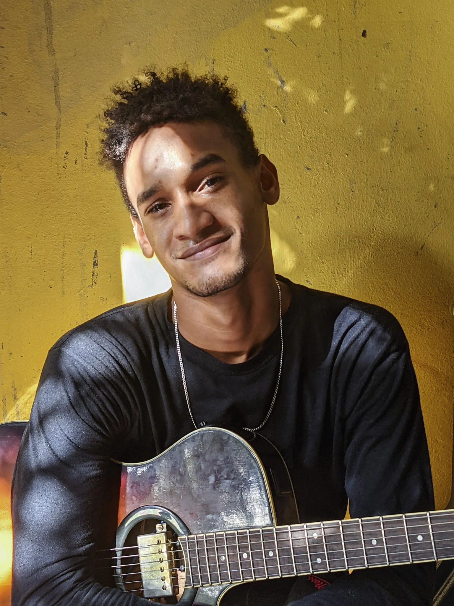 Young man with short curly hair and a black shirt smiles while holding a guitar against a bright yellow wall.