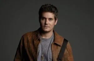 Portrait of a man with short dark hair wearing a brown leather jacket and gray tee, against a dark backdrop.