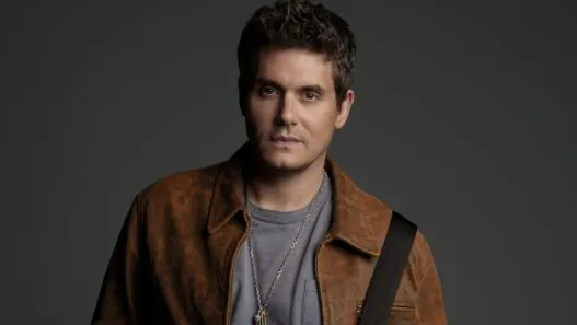 Portrait of a man with short dark hair wearing a brown leather jacket and gray tee, against a dark backdrop.
