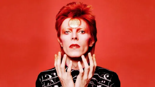 Portrait of a person with bright red hair and red lipstick against a red background, wearing a black studded jacket.