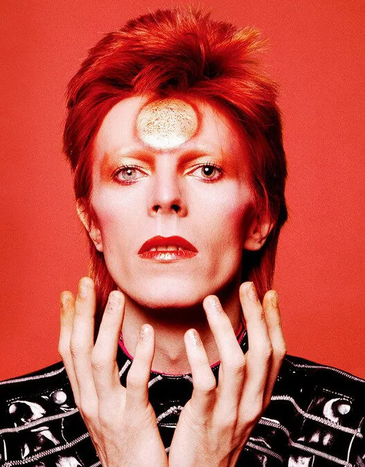 Portrait of a person with bright red hair and red lipstick against a red background, wearing a black studded jacket.