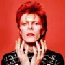Portrait of a person with bright red hair and red lipstick against a red background, wearing a black studded jacket.